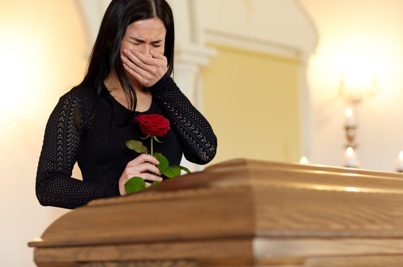 burial, people and mourning concept - crying unhappy woman with red rose and coffin at funeral in church