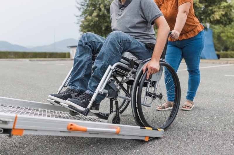 Female assistant pushing a man with disability, a wheelchair user up the car ramp into the van. Accessibility and travel concepts.