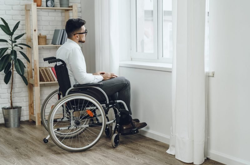 Young disabled man sitting in a wheelchair near the window at home